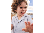 Smiling boy with curly hair wearing a light blue short-sleeved shirt, reaching out.