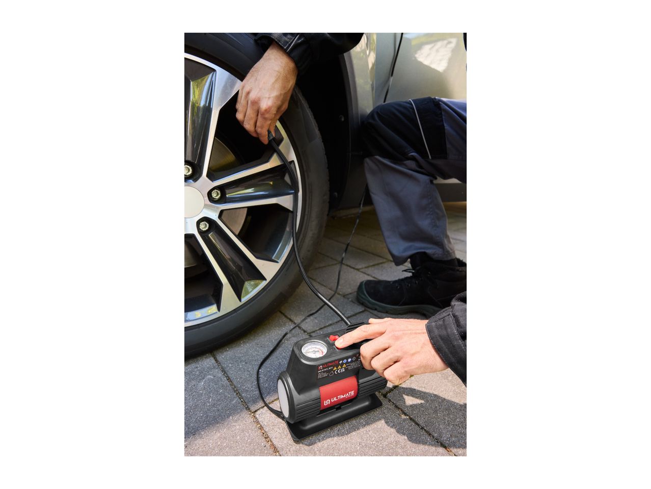 A person using a portable air compressor to inflate a car tire on a paved driveway.