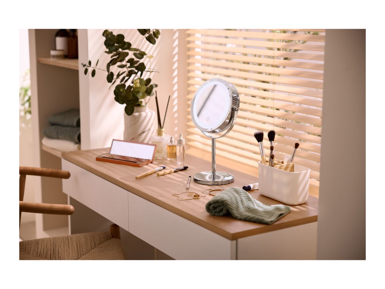 Vanity table with makeup, brushes, mirror, and a plant, next to a window with blinds.