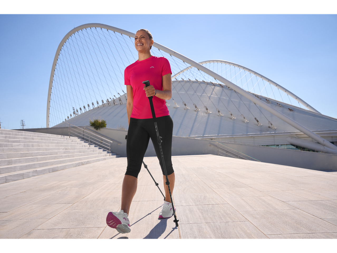 Жена в розова функционална тениска Crivit и черен клин, с щеки за скандинавско ходене.