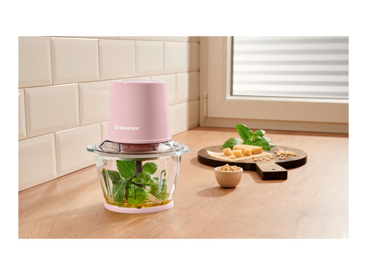 Pink food chopper with basil and oil, next to cheese and pine nuts on a wooden board.