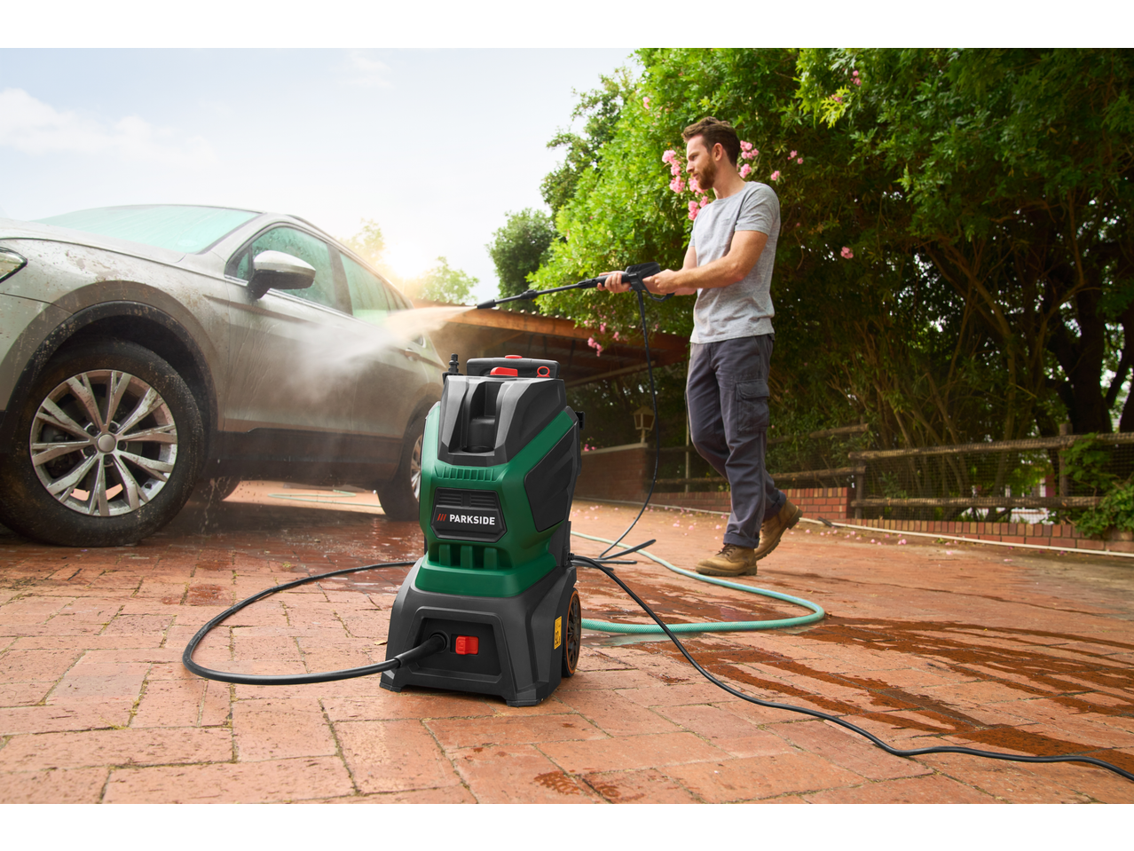 Man using a PARKSIDE® 1800W Pressure Washer to clean a dirty car on a brick patio.