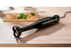 A black Silvercrest hand blender rests on a light wood surface near a cutting board with basil and cheese.