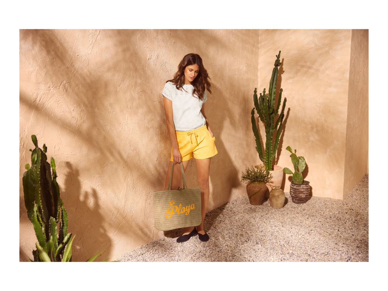 Woman in white top and yellow shorts holding a straw bag with 'Playa' text, surrounded by cacti.