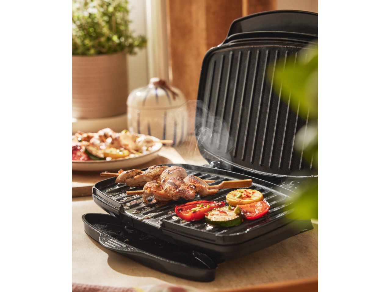 A black electric grill with chicken skewers and vegetables, with a plant in the background.
