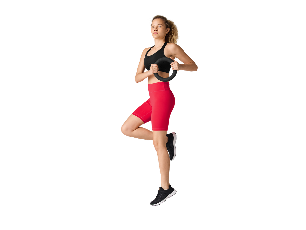 Žena u CRIVIT sportskom grudnjaku i crvenom šortsu, drži Pilates prsten, balansira na jednoj nozi.