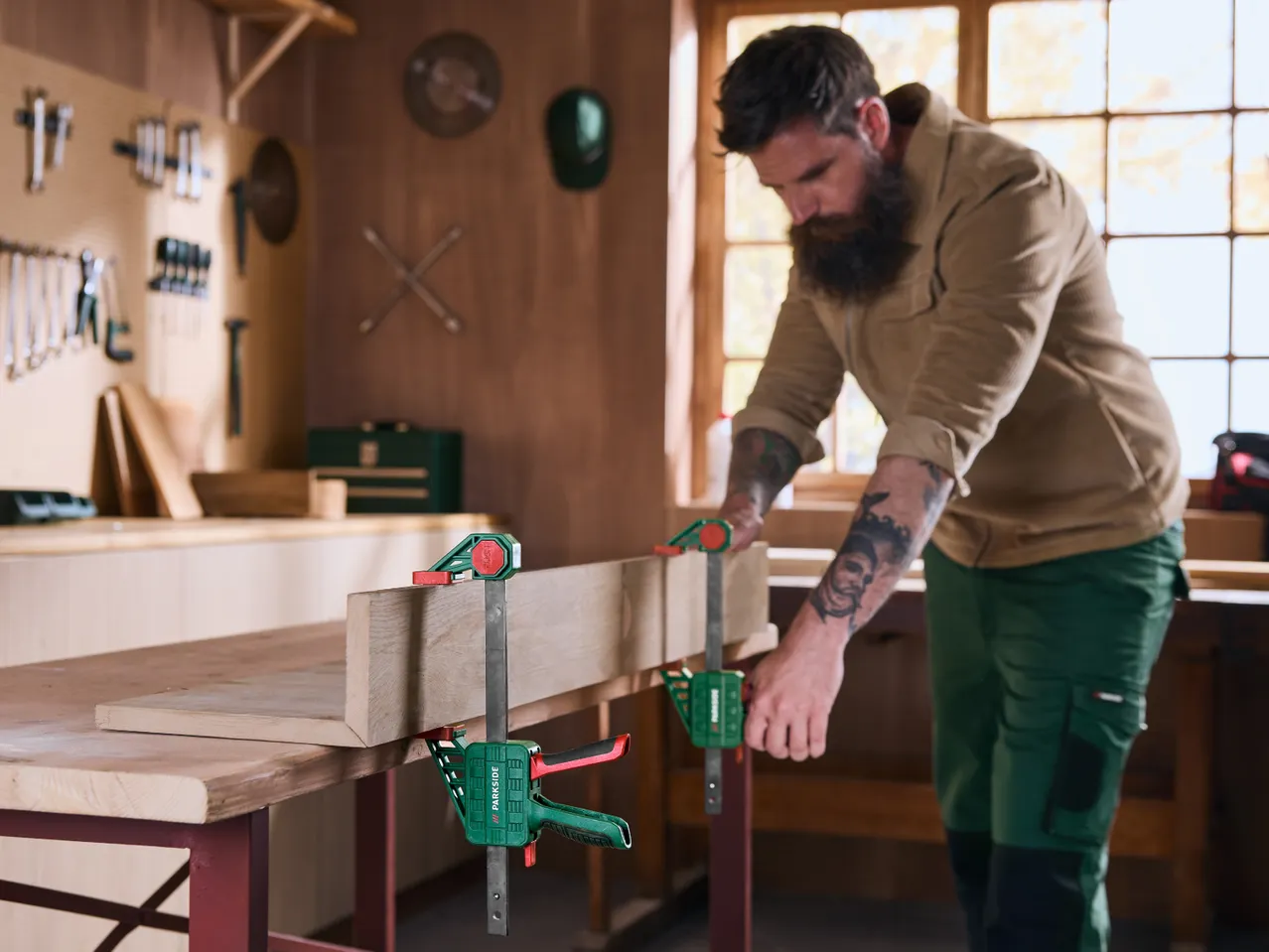 Man in a workshop using clamps to secure wood on a workbench, surrounded by tools.