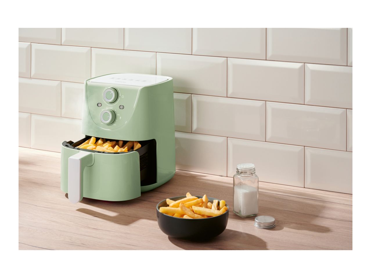 A light green air fryer with fries inside and a bowl of fries next to it on a wooden counter.
