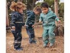 Three children in rain suits and boots splash in muddy puddles.