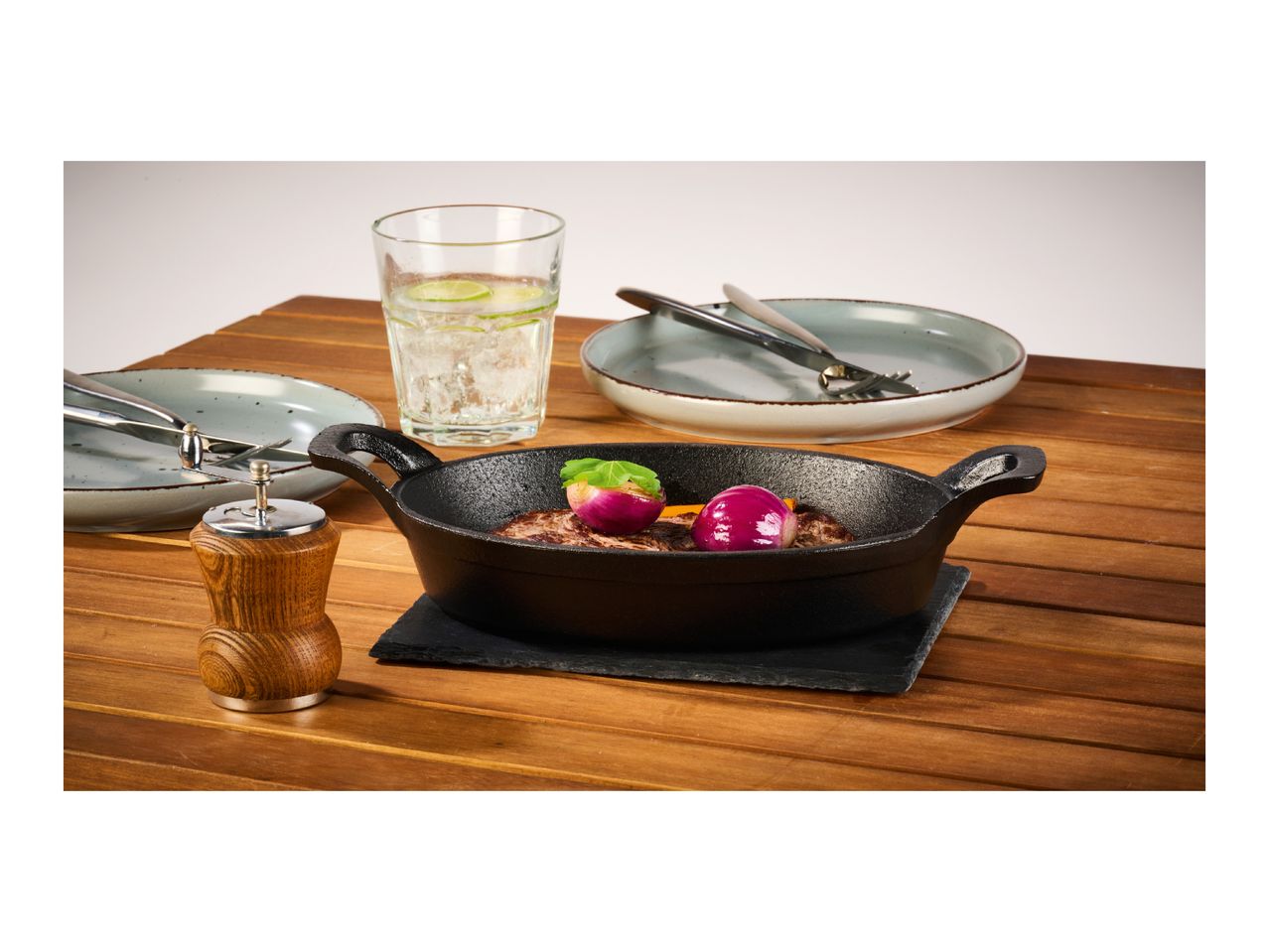 Cast iron serving pan with steak and red onions, accompanied by plates, cutlery, and a drink.