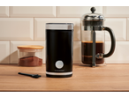 Moedor de café preto, recipiente com café moído e prensa francesa em mesa de madeira.