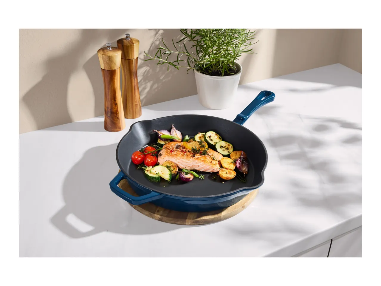 Cast iron pan with salmon and vegetables, next to salt and pepper mills and a potted herb.