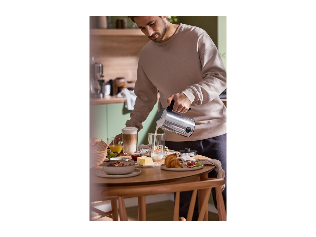 Man pouring milk from a frother into a glass, with breakfast items on a wooden table.