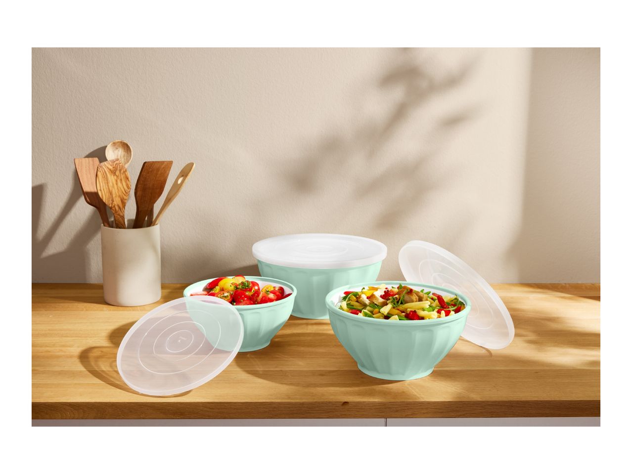 Mint green bowls with food and lids on a wooden counter, with kitchen utensils in a holder.