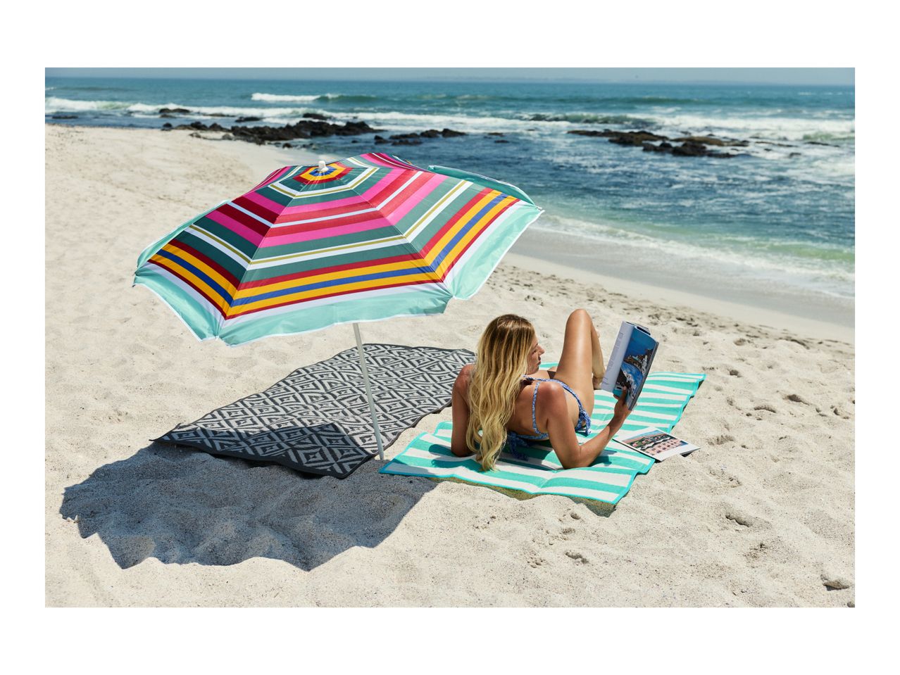 Woman reading on a striped beach mat under a colorful umbrella by the ocean.