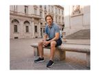 Man in a blue polo shirt and shorts sitting on a stone bench in a city square.
