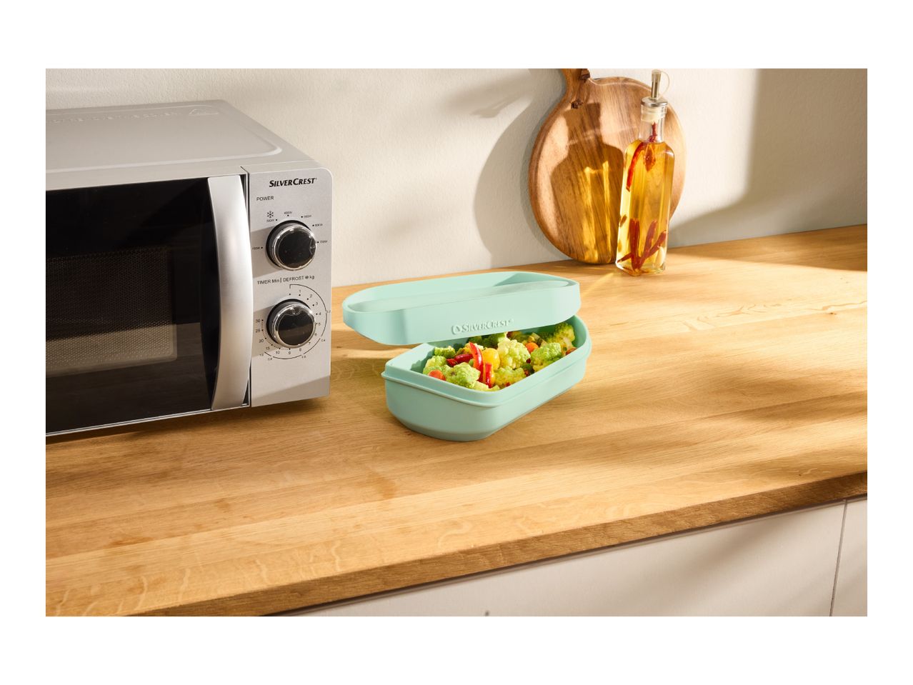 A microwave and a light green food container with vegetables on a wooden kitchen counter.