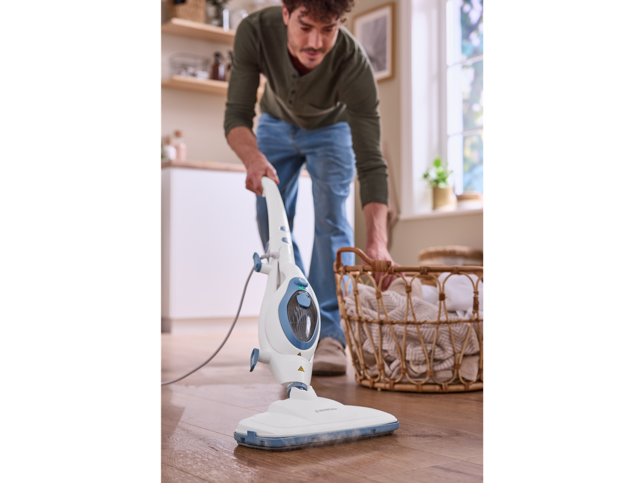 Homem a limpar o chão com o limpador a vapor Silvercrest® 2 em 1, ao lado de um cesto de roupa.