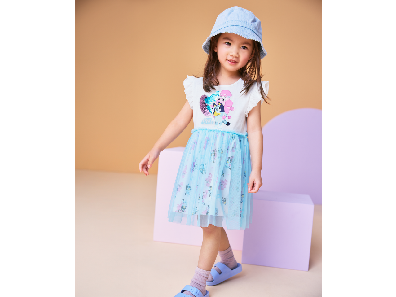 Young girl in a white and blue tulle dress with cartoon characters, wearing a denim bucket hat and purple sandals.