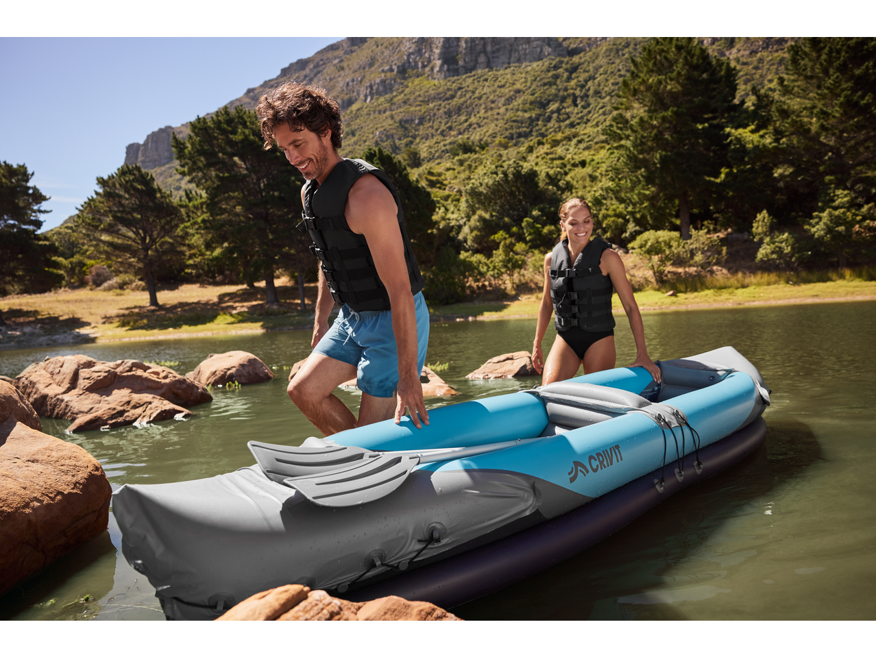 Two people in life vests getting into an inflatable kayak on a lake.