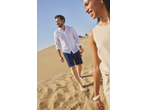 Man in white linen shirt and navy shorts walking barefoot on sand dunes with a woman.