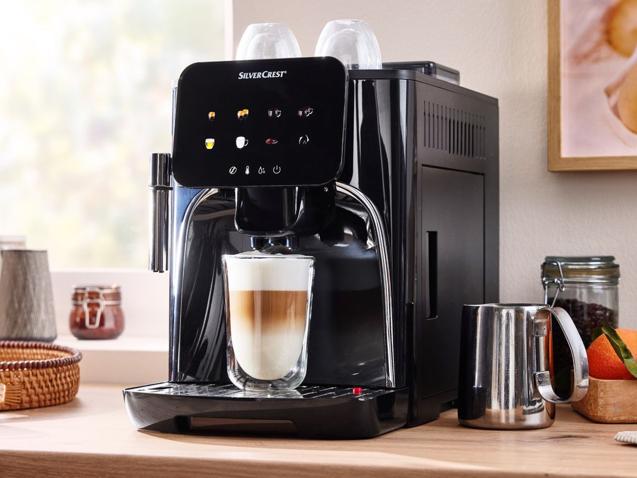 A SilverCrest coffee machine with a latte in a glass, a metal pitcher, and coffee beans.