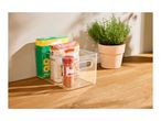 Clear storage box with pasta, spices, and a potted herb plant on a wooden surface.