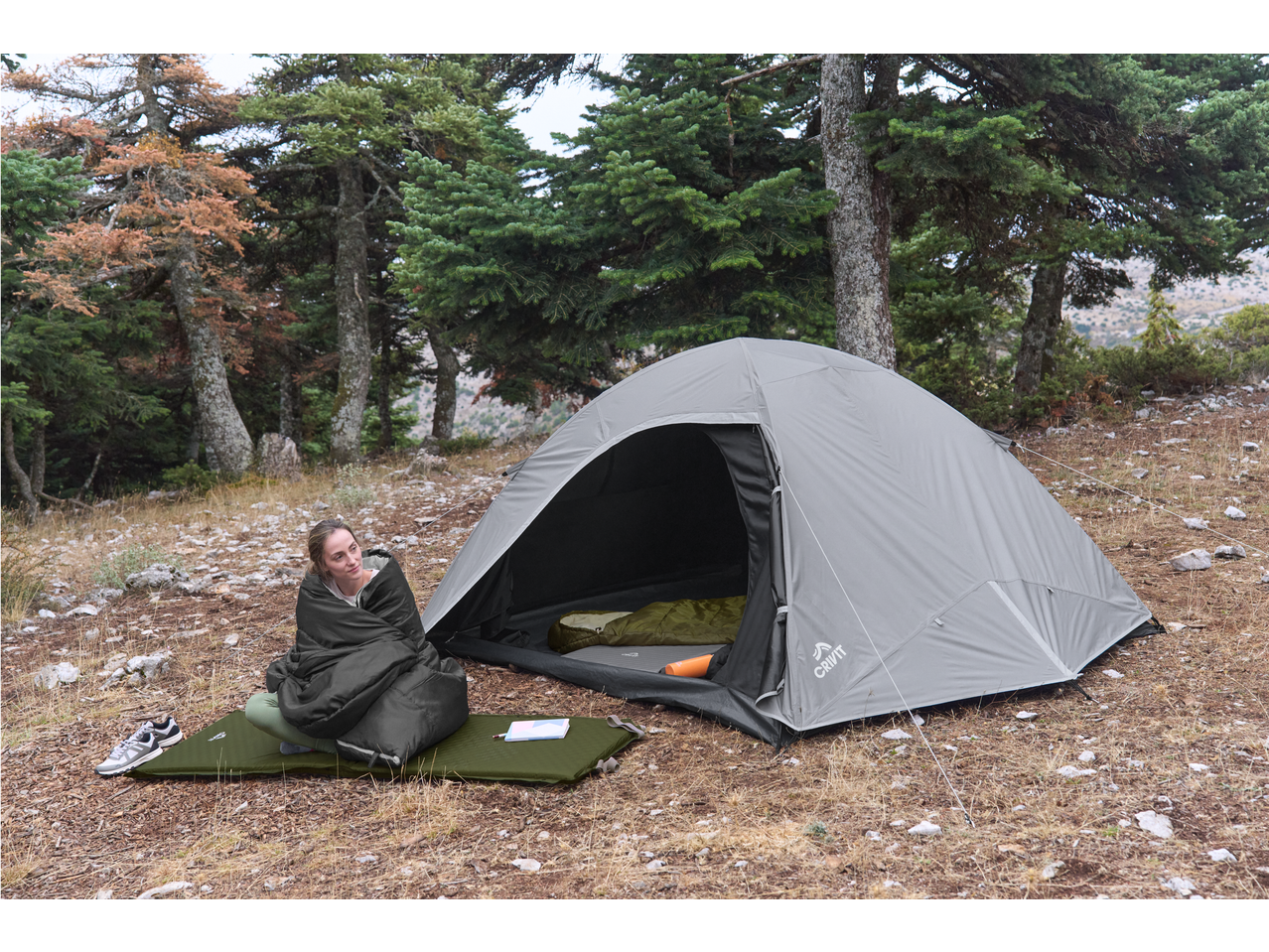 A woman in a sleeping bag sits outside a tent with camping gear in a forest.