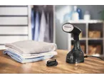 Vaporizador de roupa preto com escova e roupas dobradas numa mesa de madeira.