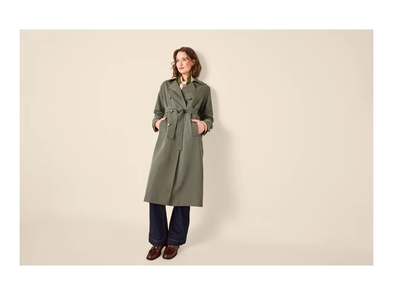 Woman in a long olive green trench coat, dark jeans, and brown loafers.