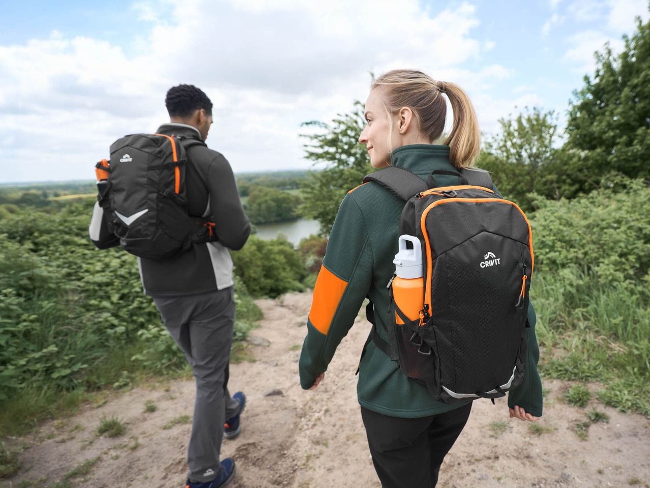 Two hikers with black and orange backpacks walk on a dirt path overlooking a lake.