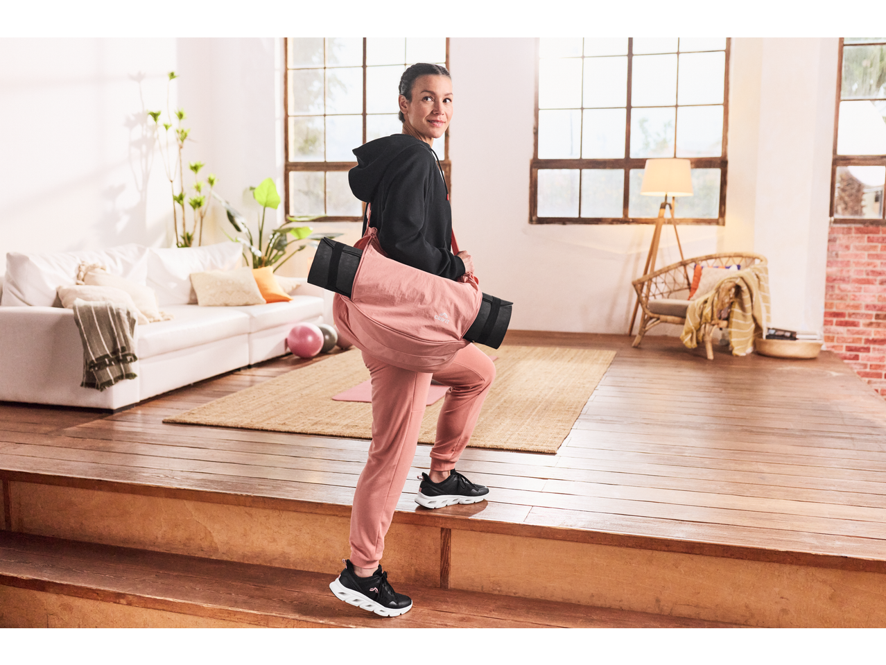 Woman in CRIVIT joggers and hoodie, carrying a yoga mat, walking up wooden steps.