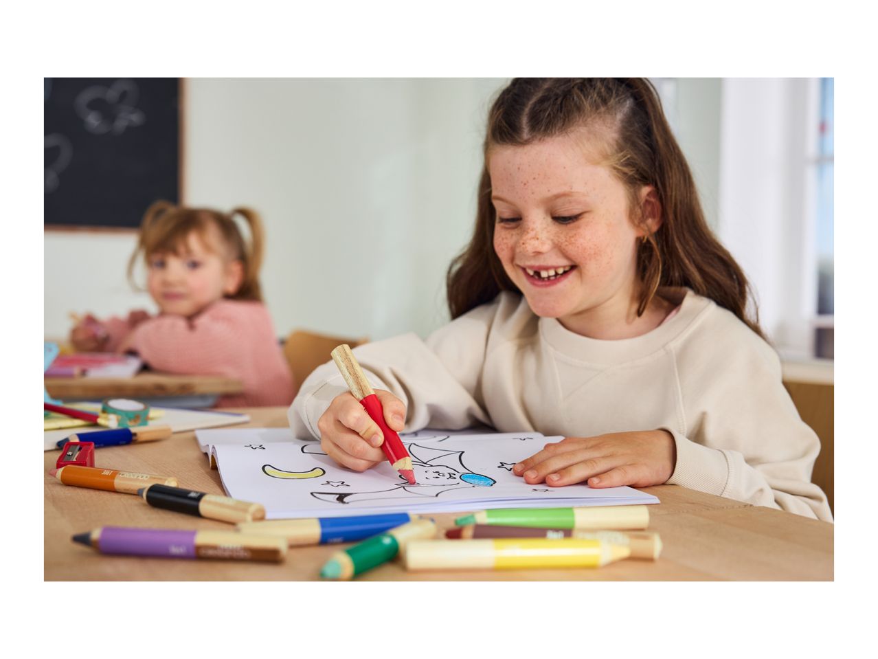 Two smiling girls coloring with chunky pencils and crayons at a wooden desk.