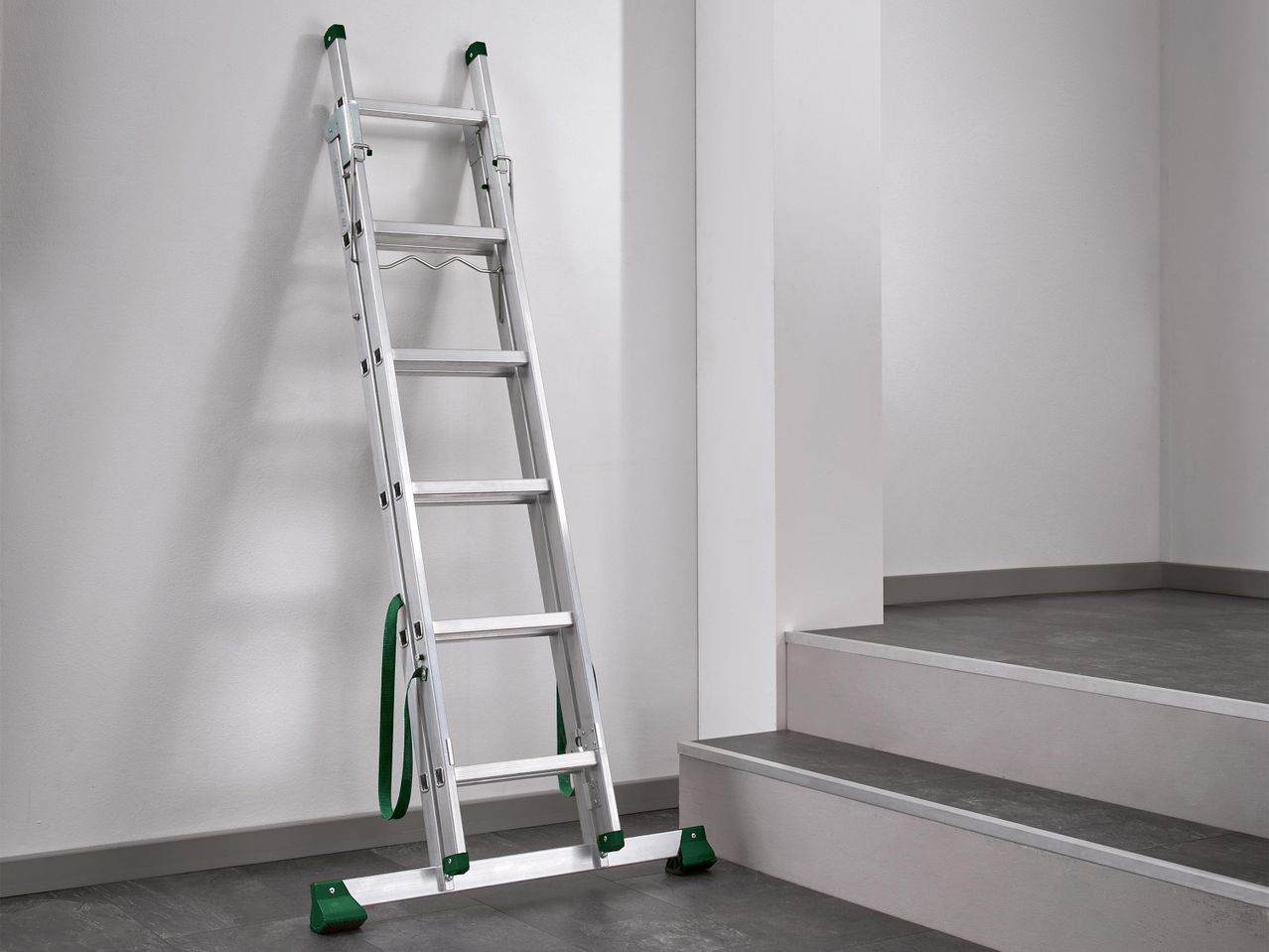 Többcélú alumínium létra zöld lábakkal és hevederekkel, falnak támasztva egy lépcső mellett.