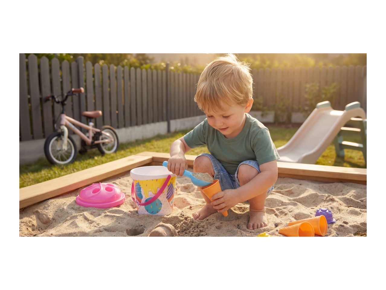 Toddler playing in a sandbox with a bucket, shovel, and sand molds.