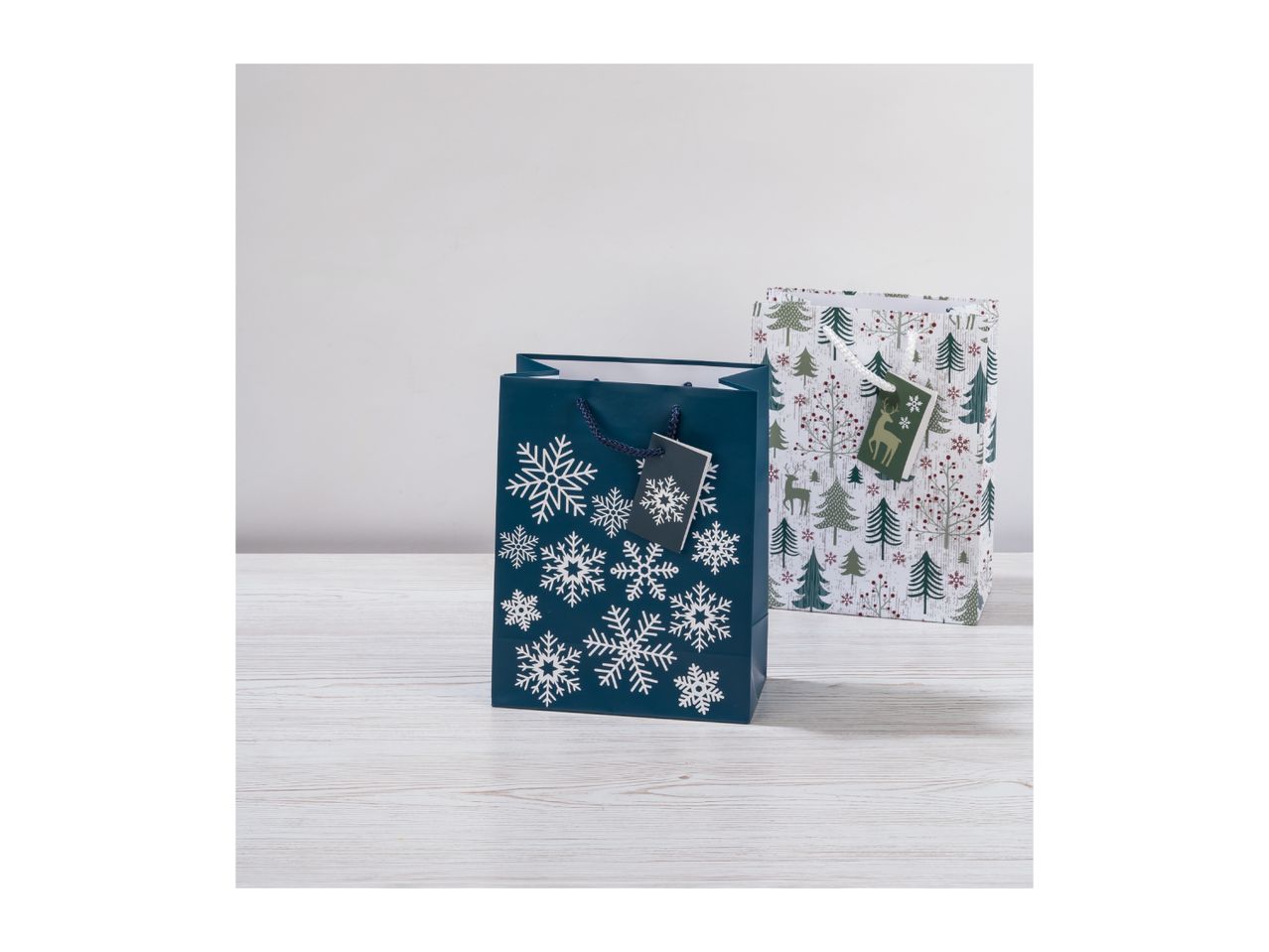 Two Christmas gift bags, one blue with snowflakes and one white with trees and deer.