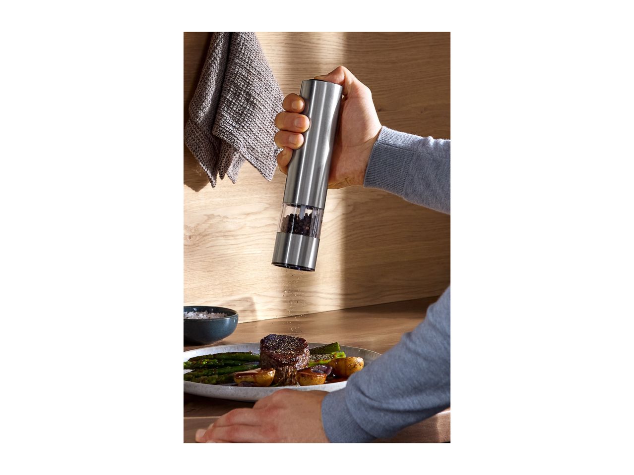Hand grinding pepper onto a steak and asparagus dish with a modern electric mill.