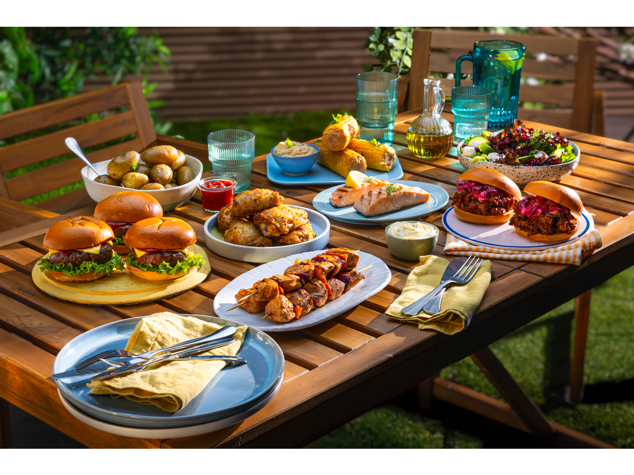 A wooden table laden with grilled burgers, chicken wings, salmon, kebabs, and salads.