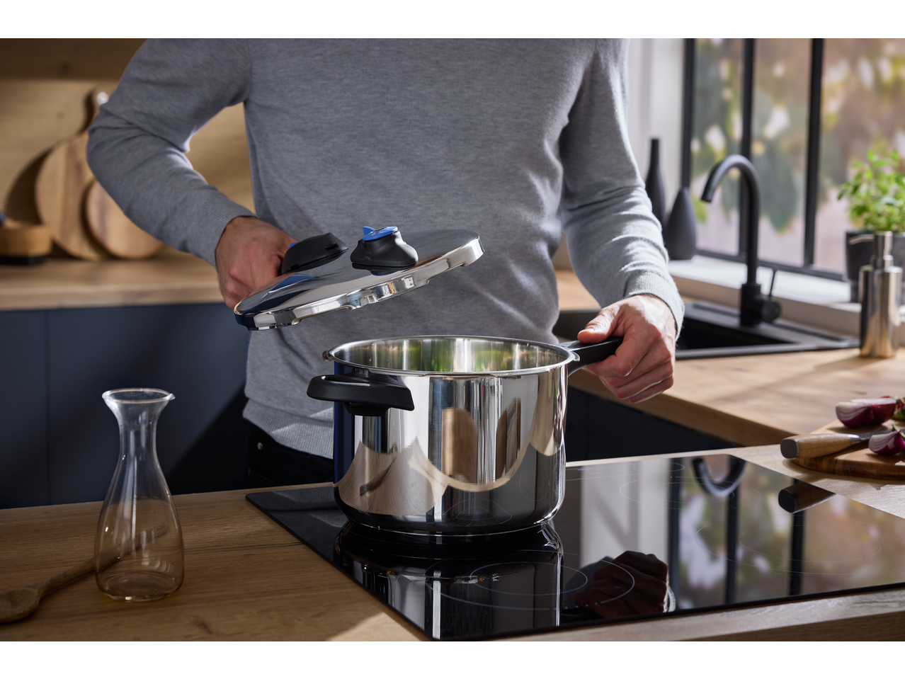 Man in a modern kitchen opening a stainless steel pressure cooker on an induction hob.