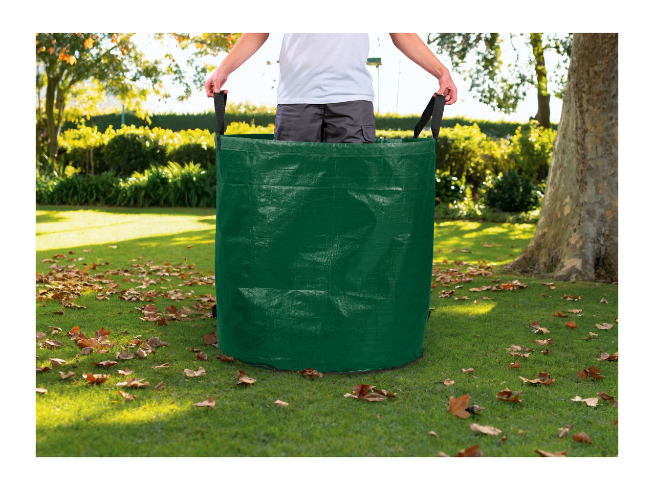 A person holding a large green garden waste bag on a lawn with fallen leaves.