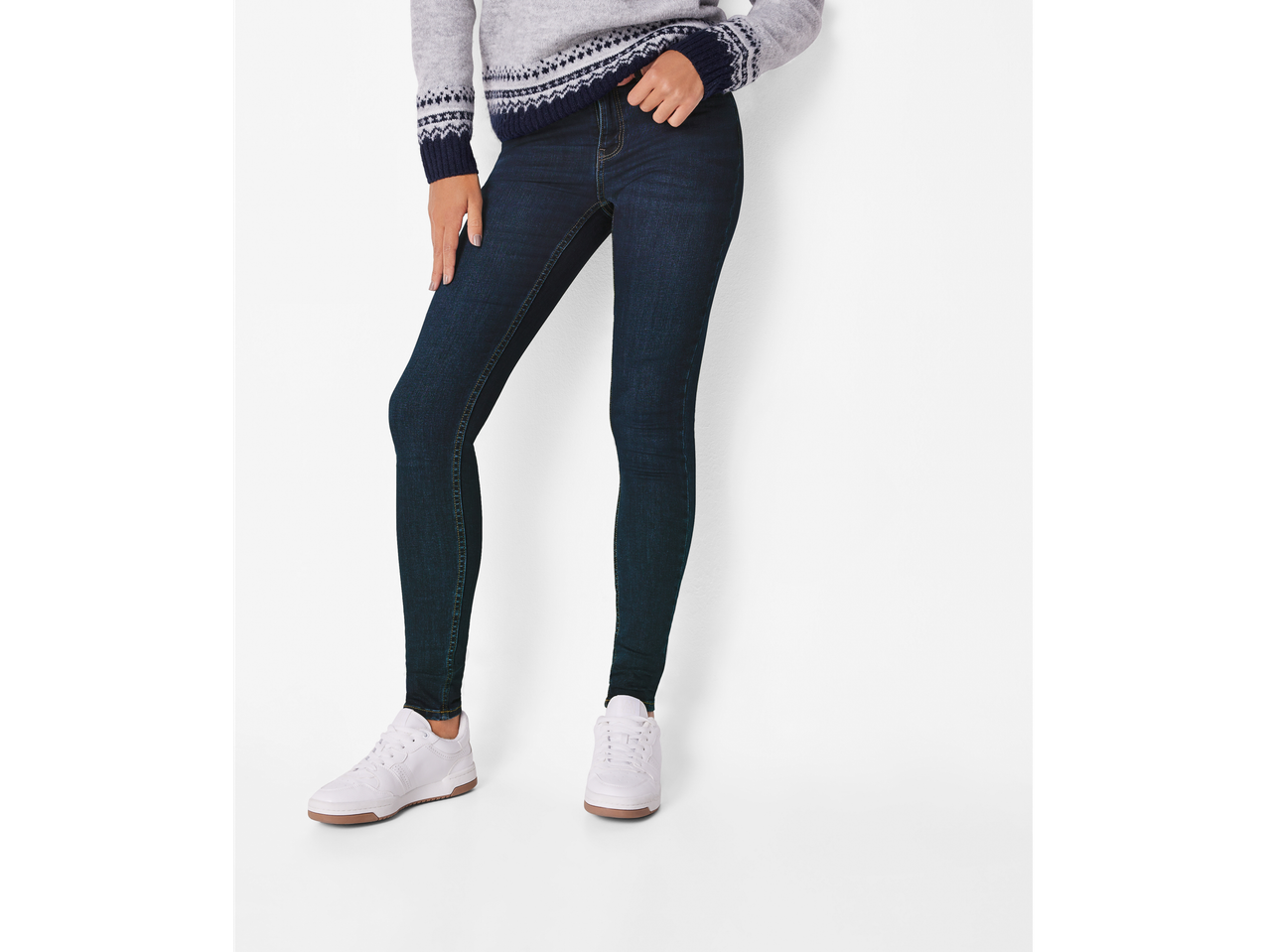 A woman wearing dark blue super skinny fit jeans and a gray patterned sweater with white sneakers.