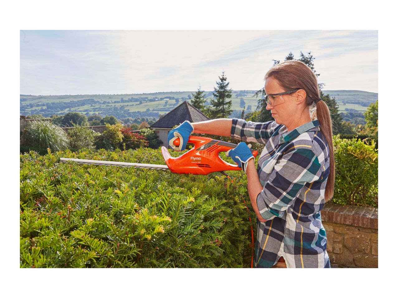 Woman trimming a hedge with an electric hedge trimmer in a garden with a scenic view.