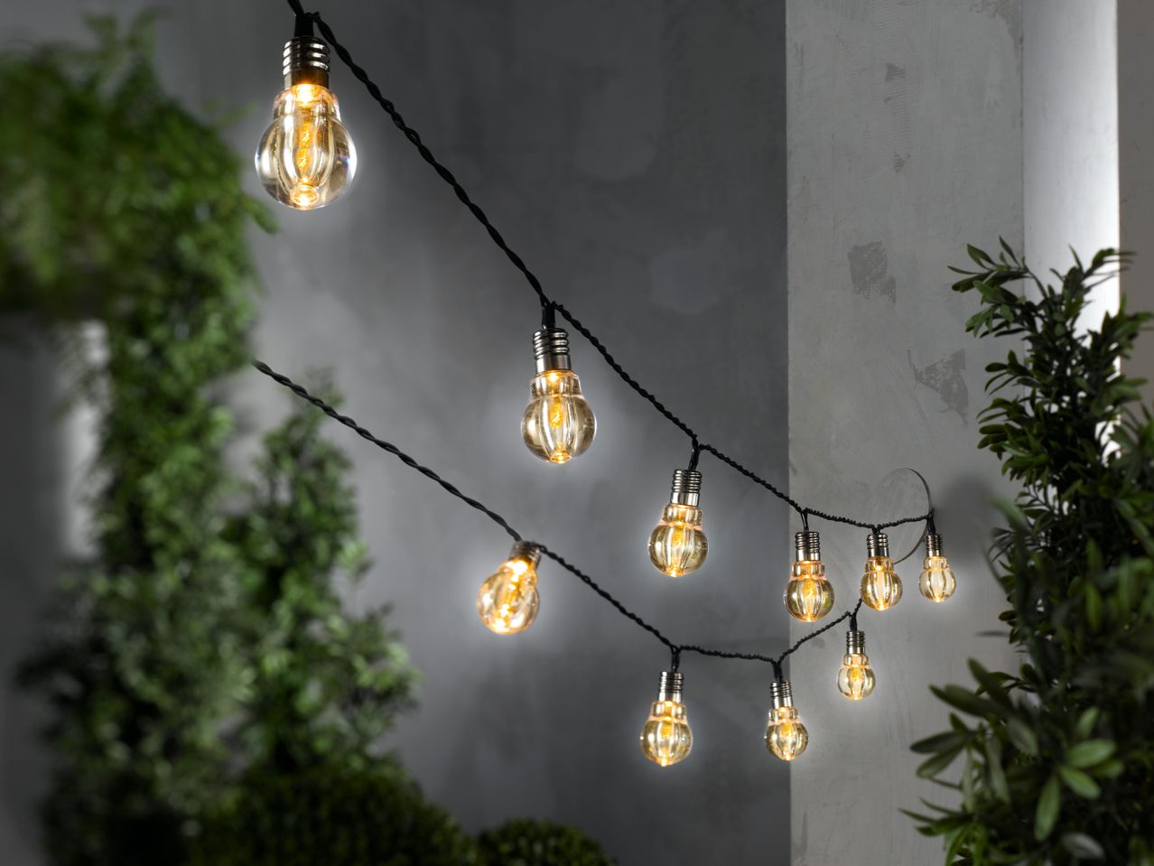 Струна с LED крушки с форма на електрическа крушка, окачена на стена, сред зелени растения.