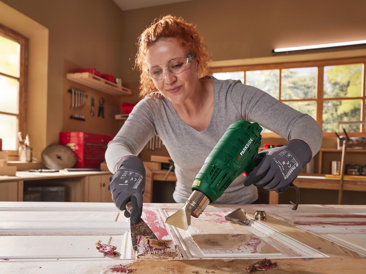 Nő Parkside hőlégfúvóval és kaparóval festéket távolít el egy ajtóról.
