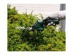 A person wearing gloves uses a cordless hedge trimmer to prune a green bush.