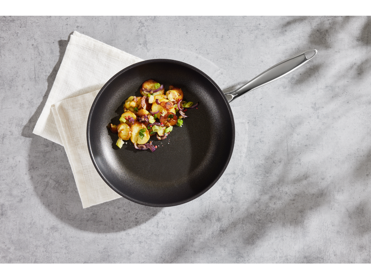 Felii de cartofi prăjiți cu ceapă și condimente într-o tigaie neagră pe un fundal gri.