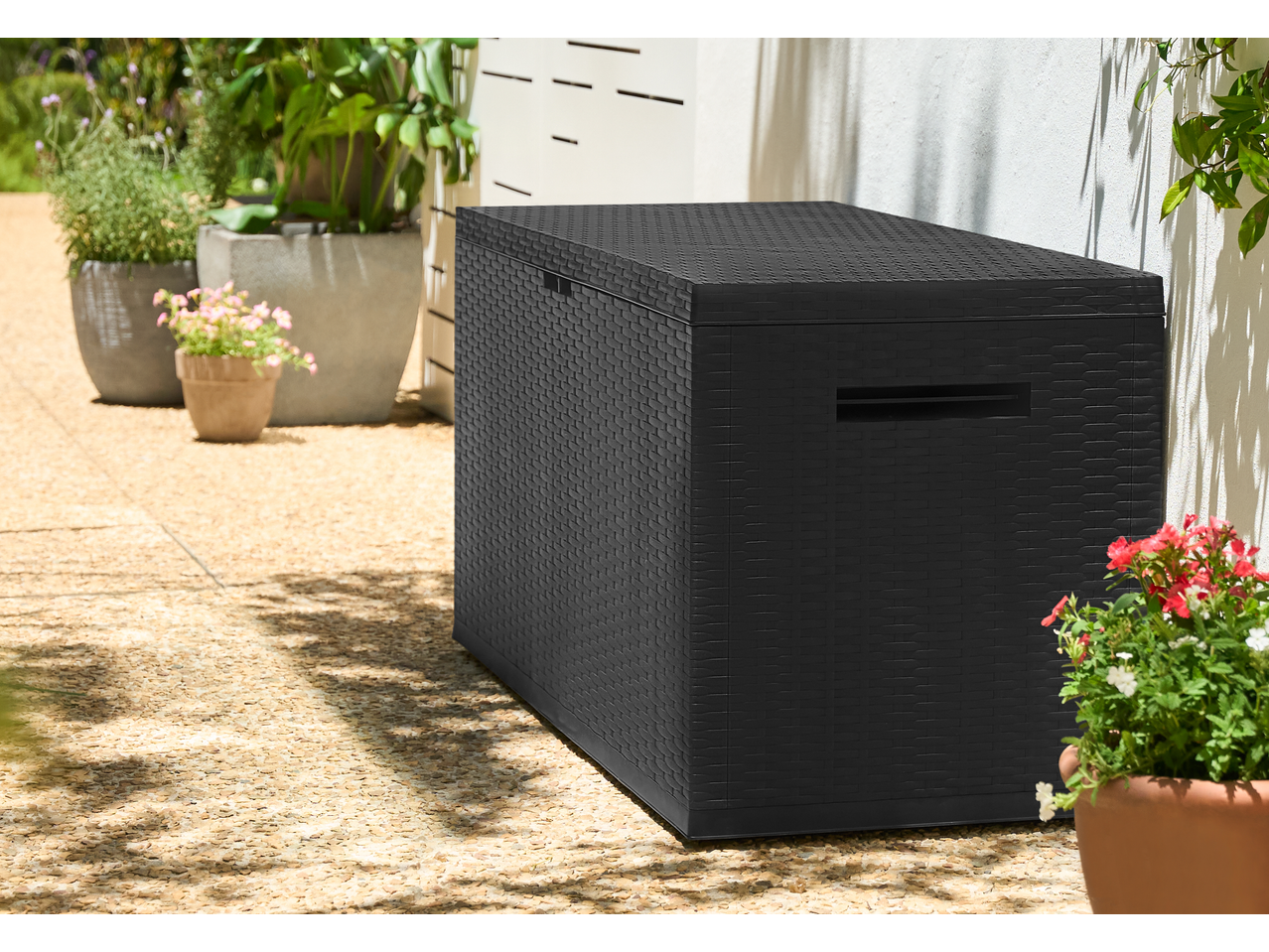 Black rattan-style outdoor storage box on a patio with potted plants.