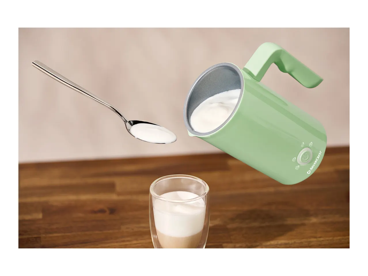 A SilverCrest milk frother pouring frothed milk into a glass with a spoon of milk floating nearby.