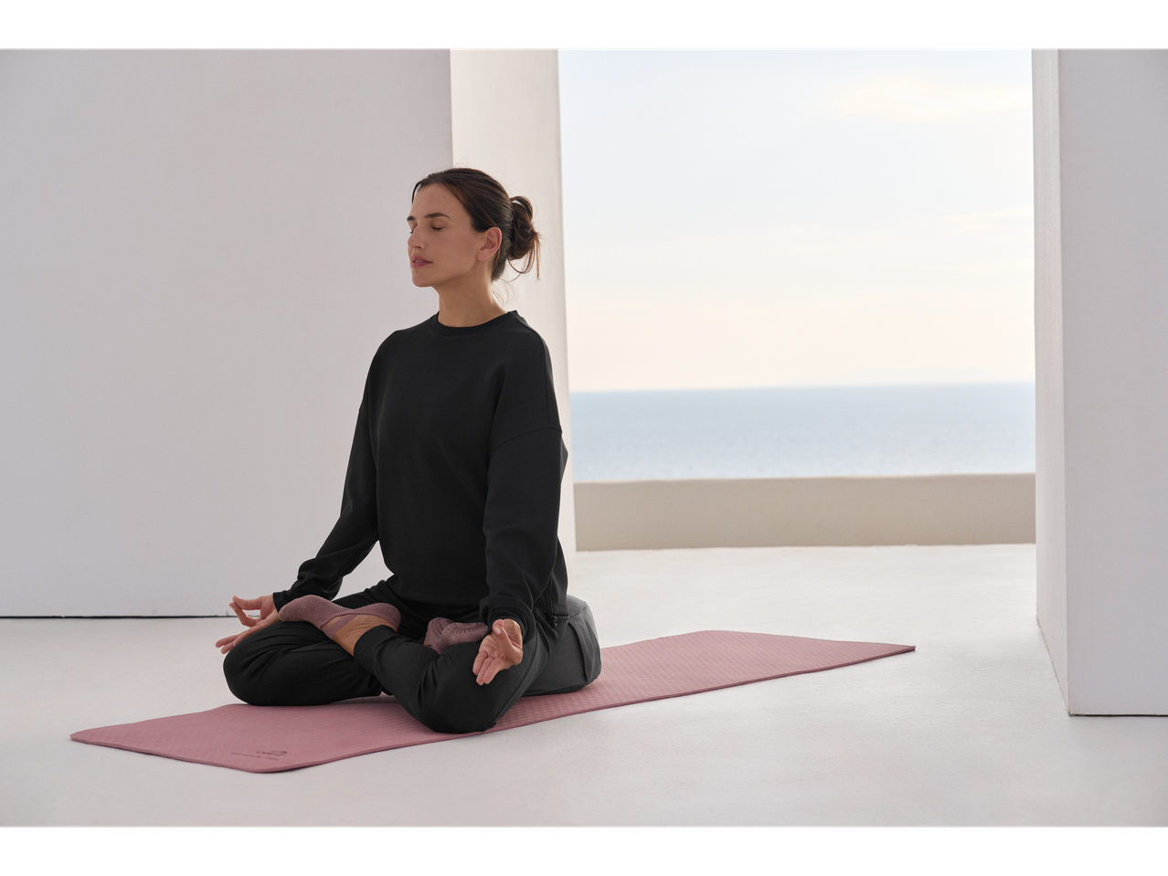 Žena meditira na prostirci za jogu, obučena u crni duks i pantalone.