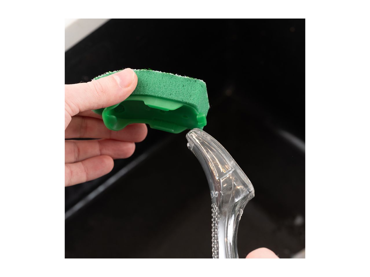 Hand holding a green dishwashing sponge with a clear soap dispenser in a sink.
